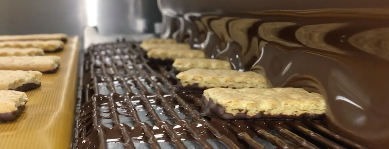 Biscuits travel along the wire mesh conveyor belt of a BETEC chocolate enrobing machine and pass through a curtain of tempered milk chocolate, creating an even chocolate coating during the enrobing process.